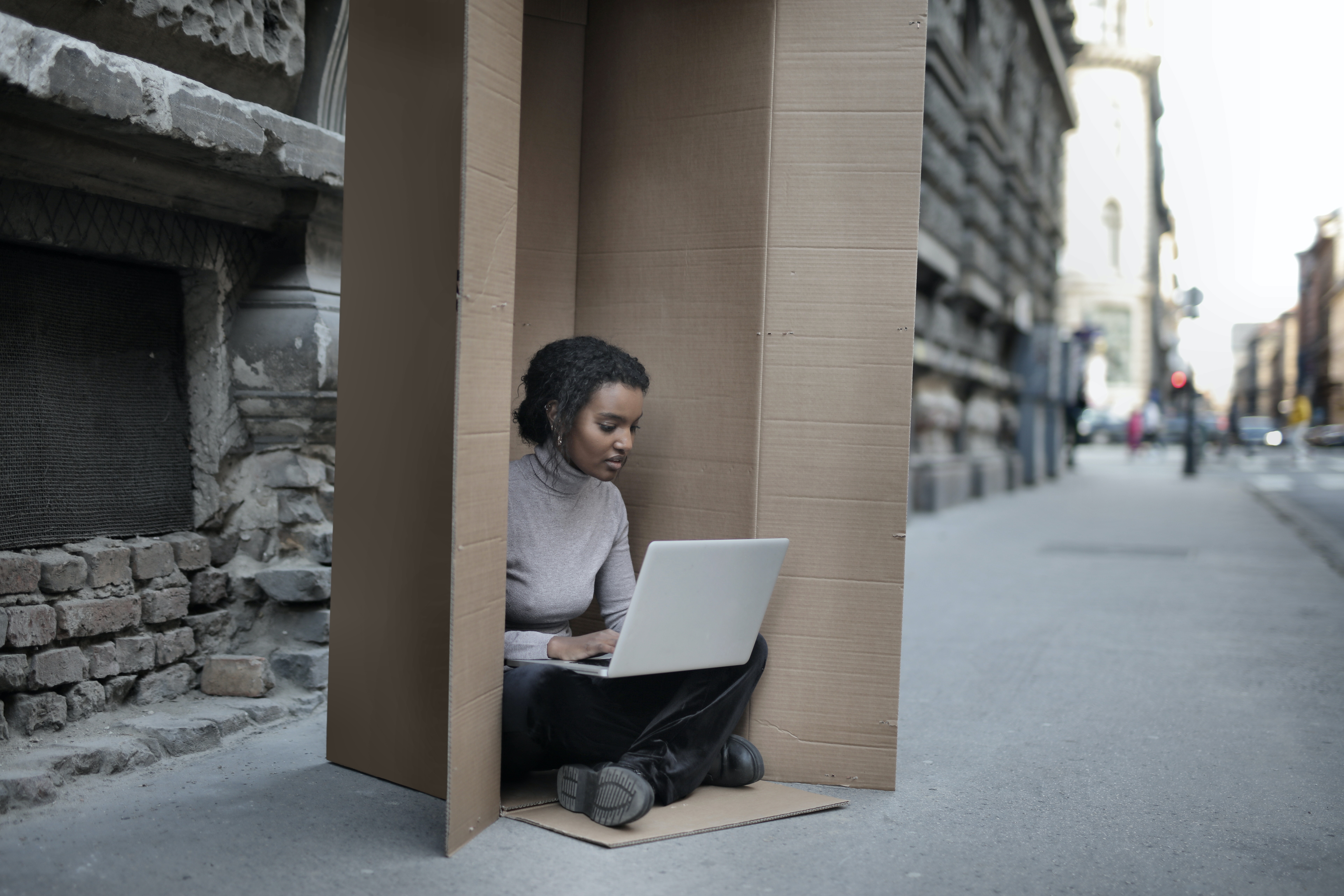 Kobieta z laptopem siedząca w kartonowym pudle na chodniku